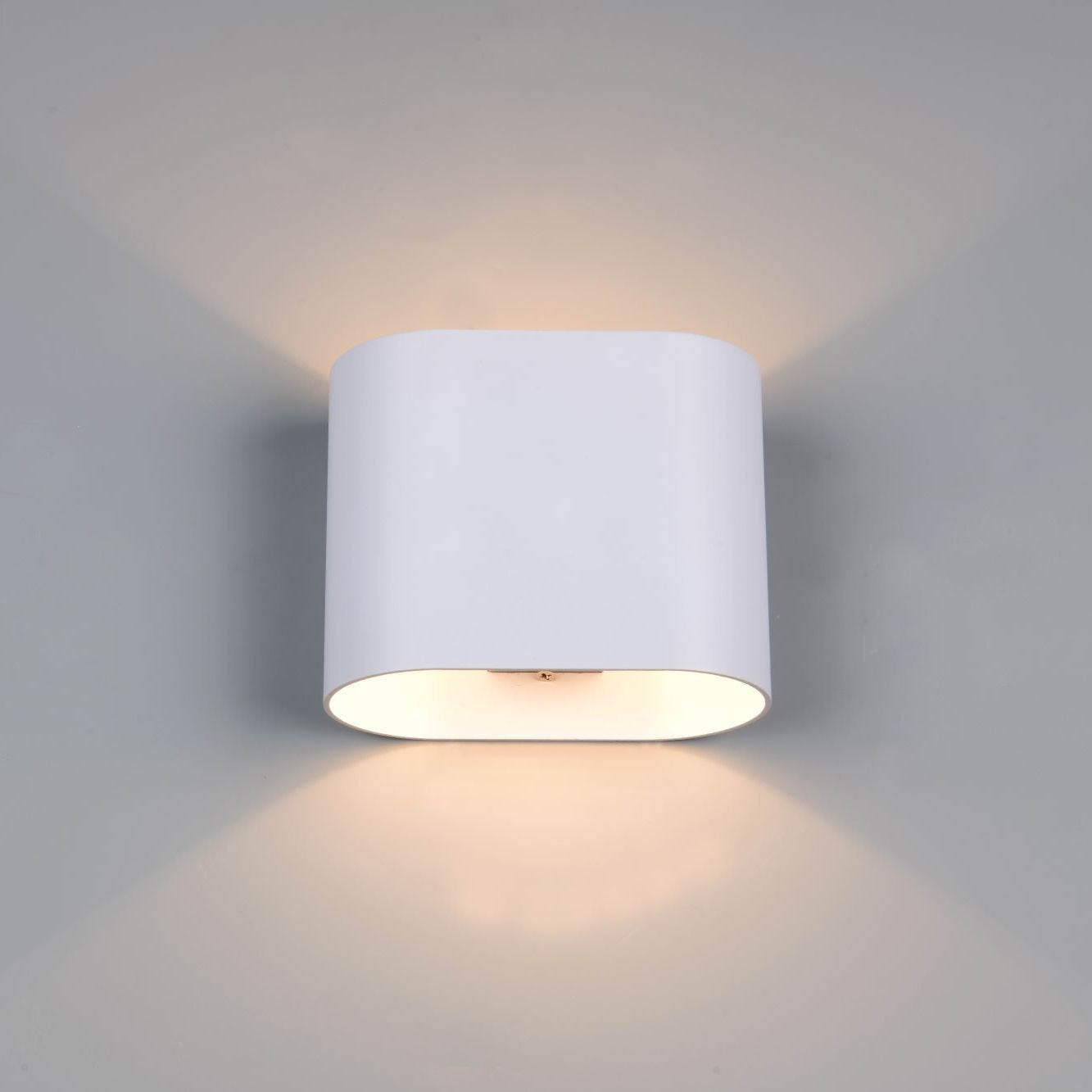 A modern cylindrical wall sconce light fixture in matte white, mounted on a light gray wall and casting a warm glow upwards and downwards.