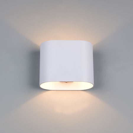 A modern cylindrical wall sconce light fixture in matte white, mounted on a light gray wall and casting a warm glow upwards and downwards.