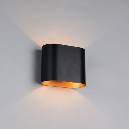 A modern black wall sconce light fixture with a warm gold interior, mounted on a light gray wall and casting a warm glow upwards and downwards.