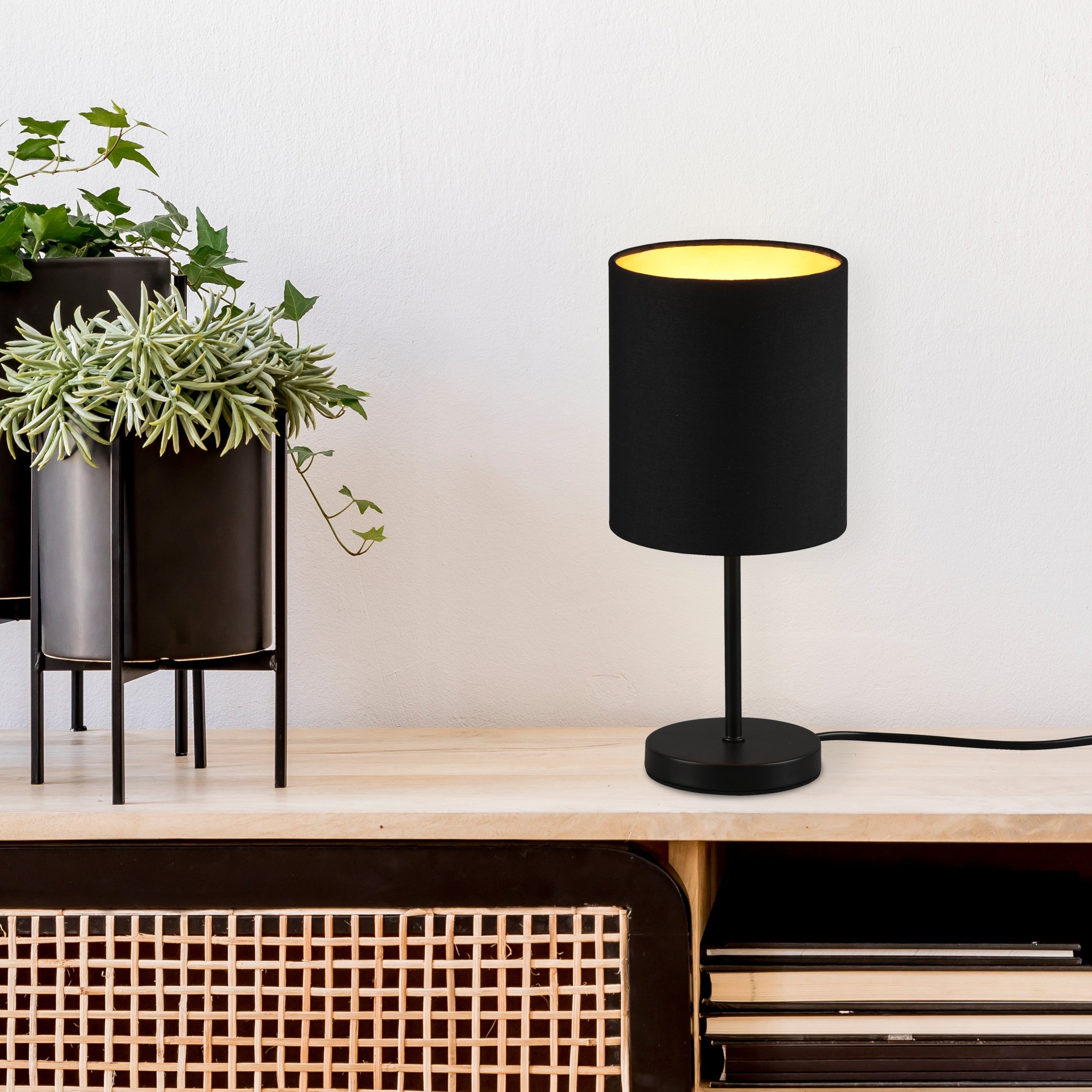 Black table lamp with a yellow interior on a wooden surface next to potted plants.