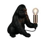 A gorilla-shaped table lamp with a black textured body, holding a glowing clear bulb in a metallic gold holder, shown on a plain white background.