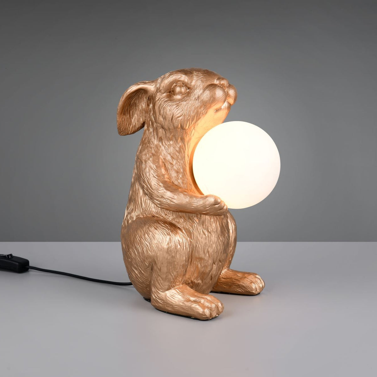 A rabbit-shaped table lamp with a metallic gold finish, holding a glowing white spherical bulb, set against a neutral gray background.