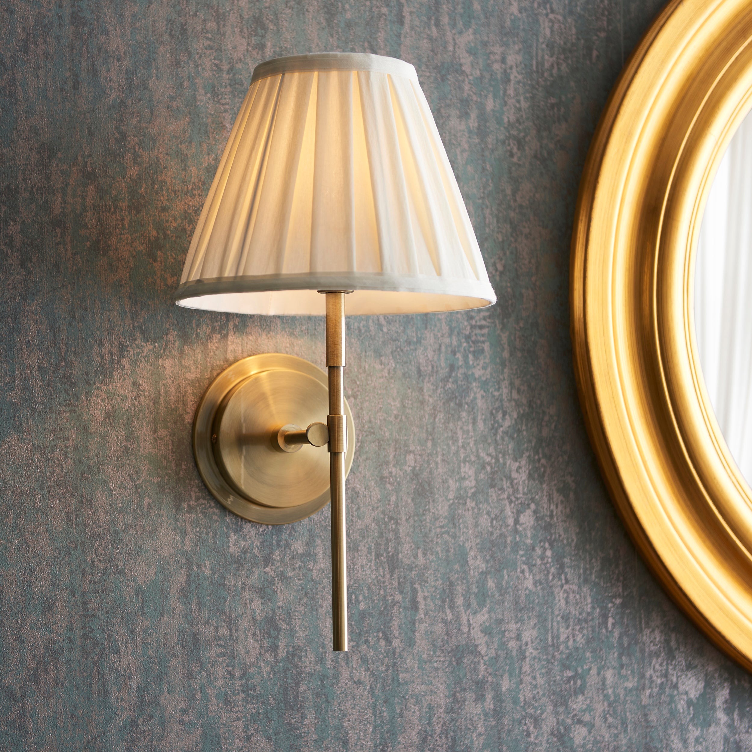 Wall-mounted lamp with pleated shade on a textured wall next to a round mirror.